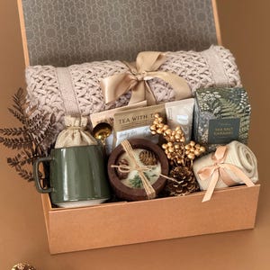 May include: A gift box containing a beige knit blanket tied with a ribbon, a dark green ceramic mug, a small burlap pouch, a wooden bowl candle with pinecones, a gold spoon, a box labeled Sea Salt Caramel, and a rolled beige cloth with a ribbon.