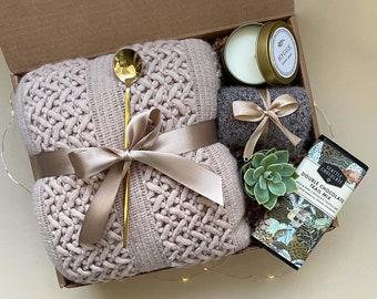 May include: A gift box filled with cozy items for a relaxing evening. It includes a pink knitted throw, a gold spoon, a small candle, a gray scarf, a succulent plant, and a bar of chocolate with the text "Seattle Chocolate Double Chocolate Trail Mix".