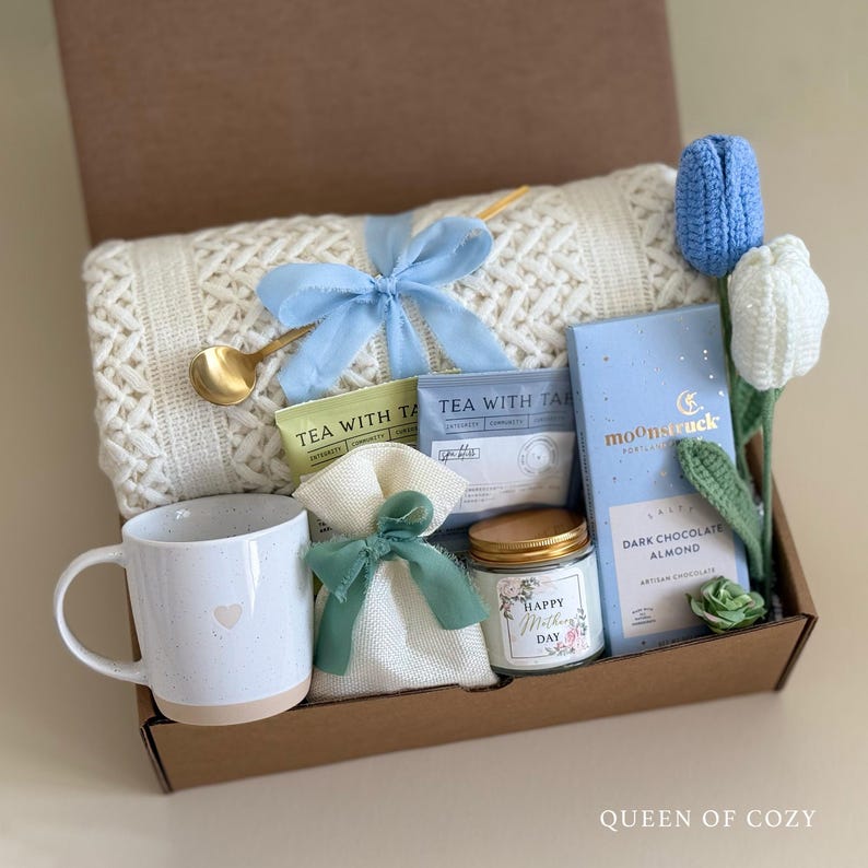 May include: A gift box filled with items for a cozy night in, including a white knitted throw blanket, a mug, a candle, a bag of tea, a bar of chocolate, and a crocheted flower.
