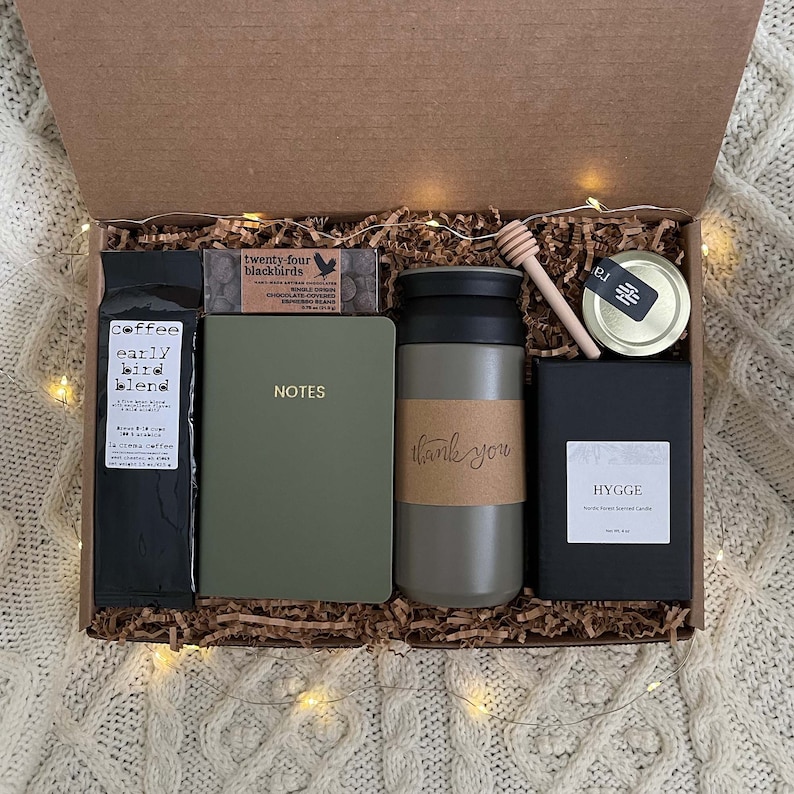 May include: A gift box filled with a variety of items, including a green notebook with the word "NOTES" on the cover, a green travel mug with the words "thank you" on it, a black coffee package labeled "coffee early bird blend", a small box of chocolate espresso beans, a small jar of honey, and a black candle labeled "HYGGE".