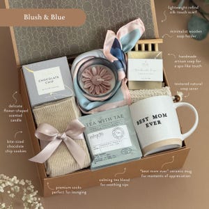 May include: A gift box labeled "Blush & Blue" filled with a variety of items. Includes a "Best Mom Ever" mug, a flower-shaped candle, a silk scarf, chocolate chip cookies, tea, socks, and soap. The box is tied with a ribbon.