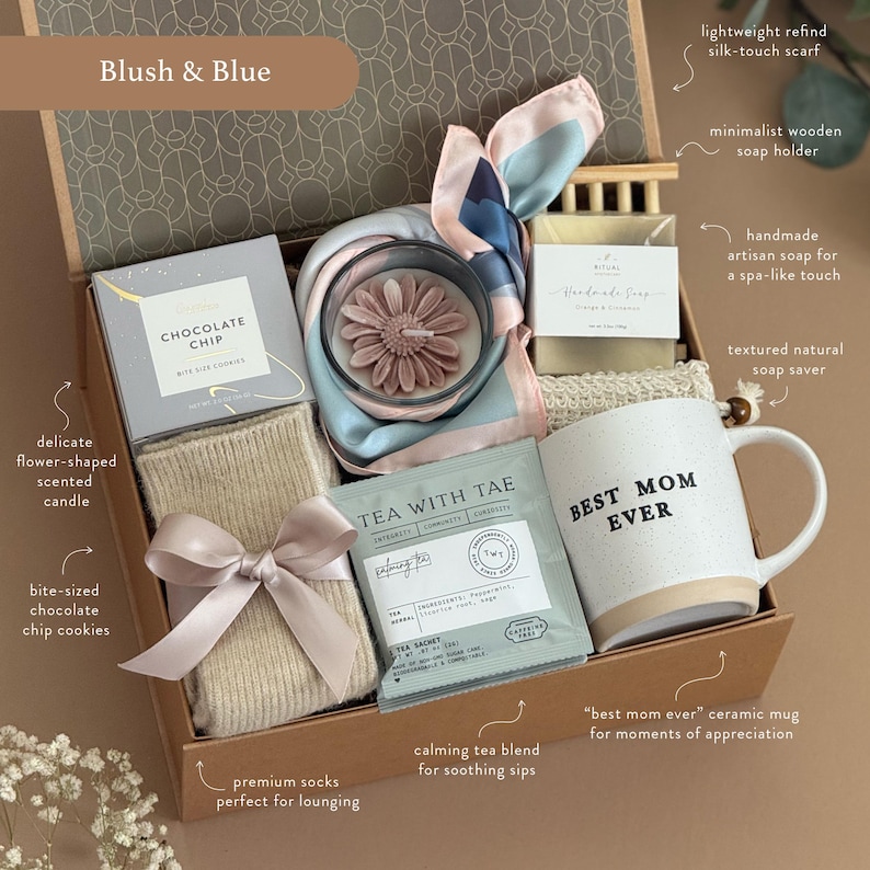 May include: A gift box labeled "Blush & Blue" filled with a variety of items. Includes a "Best Mom Ever" mug, a flower-shaped candle, a silk scarf, chocolate chip cookies, tea, socks, and soap. The box is tied with a ribbon.