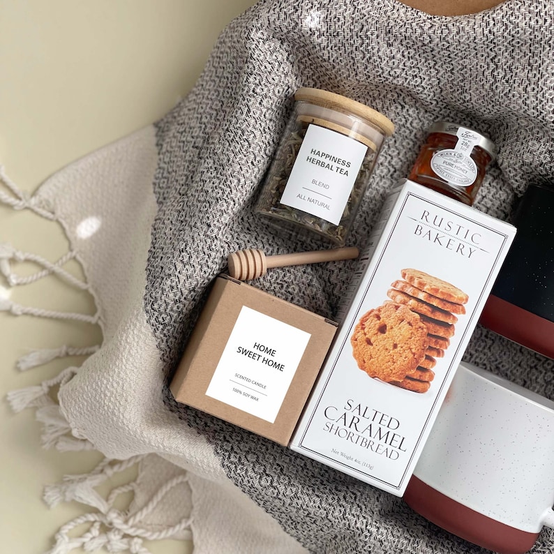 May include: A gift basket with a brown and white throw blanket, a glass jar of herbal tea labeled "Happiness Herbal Tea", a brown candle labeled "Home Sweet Home", a box of salted caramel shortbread cookies labeled "Rustic Bakery", a jar of honey, and a wooden honey dipper.