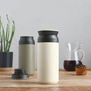 May include: Two stainless steel insulated water bottles with black lids. The bottle on the left is a muted green color, and the bottle on the right is a light cream color. The bottles are sitting on a wooden table.