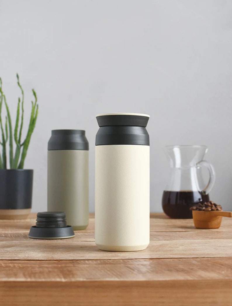 May include: Two stainless steel insulated water bottles with black lids. The bottle on the left is a muted green color, and the bottle on the right is a light cream color. The bottles are sitting on a wooden table.