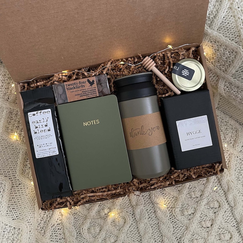May include: A brown gift box filled with a variety of items, including a green notebook with the word "NOTES" on the cover, a green travel mug with the words "thank you" on it, a black box with the word "HYGGE" on it, a small jar of honey, and a bag of coffee beans. The box is sitting on a white knitted surface.