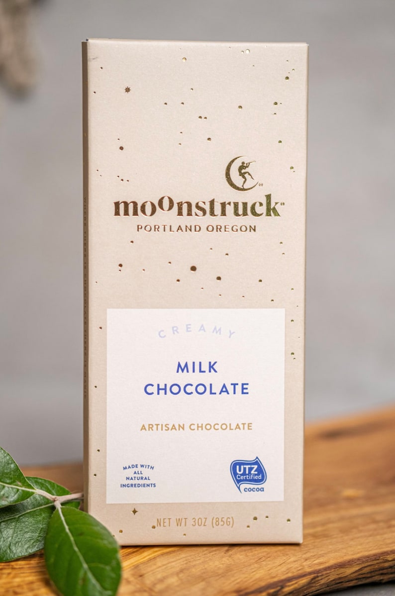 以下が含まれることがあります： 「moonstruck」と「PORTLAND OREGON」の文字が書かれた、金色と白のチョコレートバーの包装紙。包装紙には、「CREAMY MILK CHOCOLATE」と「ARTISAN CHOCOLATE」の文字も書かれている。包装紙には、金色の星模様と、その上に人が立っている小さな金色の三日月が描かれている。包装紙には、「UTZ Certified cocoa」のロゴも記載されている。バーは3オンス（85g）です。