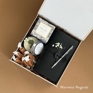 May include: A white gift box filled with a black journal, a silver pen, a small candle, a box of tea, and caramel candies. The box is decorated with white paper shreds and a small sprig of white flowers. The text "Warmest Regards" is printed on the bottom right corner of the box.