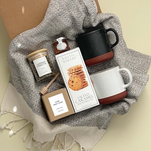 May include: A gift basket with a brown and white woven basket filled with a variety of items, including a jar of herbal tea, a jar of honey, a box of salted caramel shortbread, a box of home sweet home cookies, and two mugs. The mugs are black and white with red accents. The basket is on a light green background.