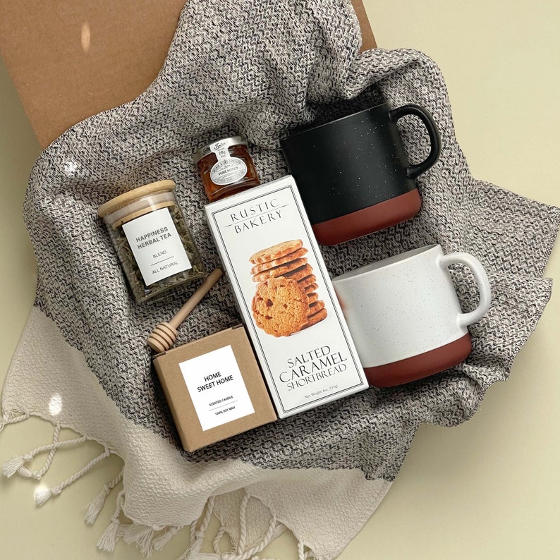 May include: A gift basket with a brown and white woven basket filled with a variety of items, including a jar of herbal tea, a jar of honey, a box of salted caramel shortbread, a box of home sweet home cookies, and two mugs. The mugs are black and white with red accents. The basket is on a light green background.