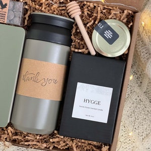 May include: A gift box filled with a gray travel mug, a black candle with the text "HYGGE Nordic Forest Scented Candle Net Wt. 4 oz", a small jar of honey, and a small box of chocolates. The box is lined with brown crinkled paper.