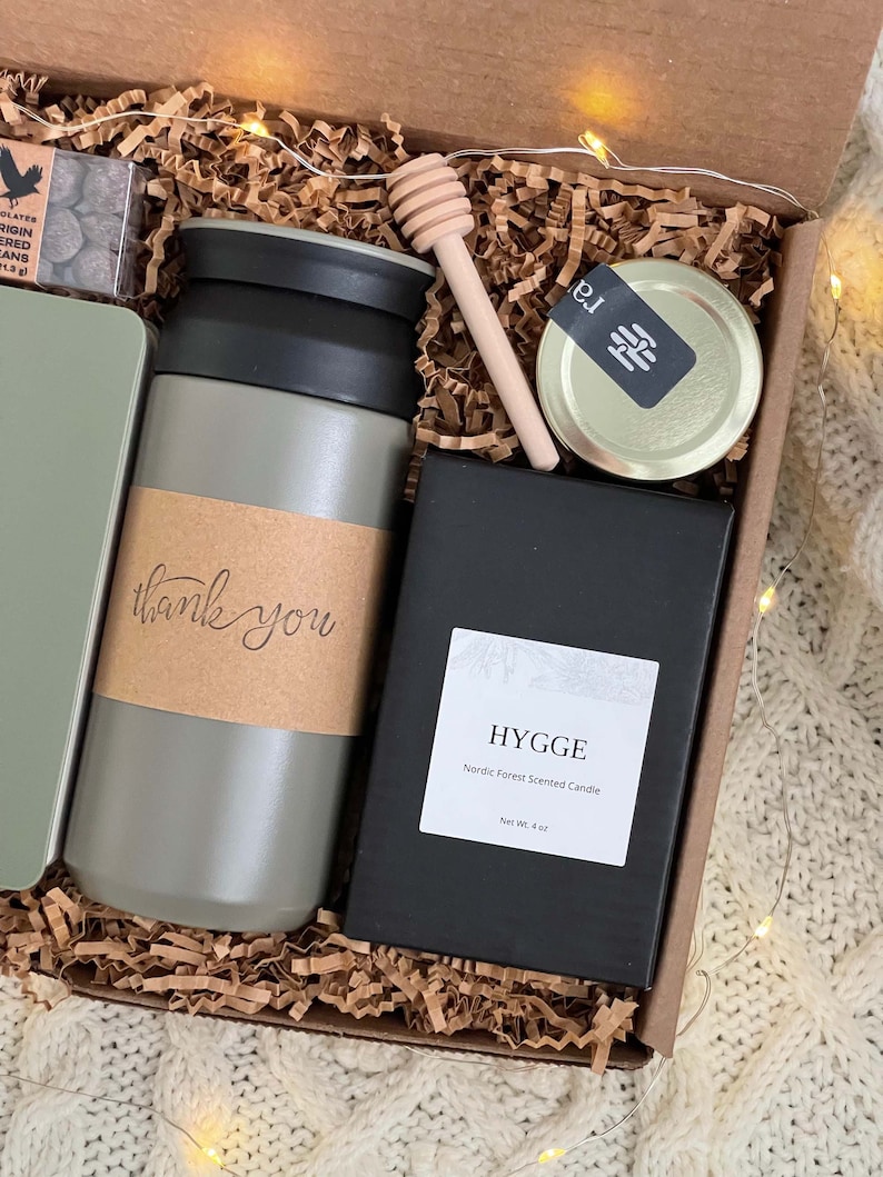 May include: A gift box filled with a gray travel mug, a black candle with the text "HYGGE Nordic Forest Scented Candle Net Wt. 4 oz", a small jar of honey, and a small box of chocolates. The box is lined with brown crinkled paper.