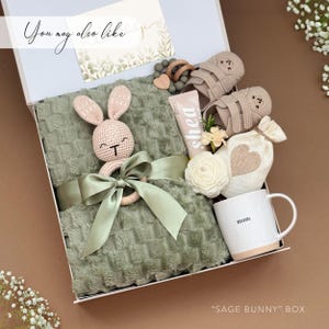 May include: A gift box with a sage green blanket, a crochet bunny rattle, a tube of shea lotion, a pair of baby shoes, a mug that says "mom", and a small bag with a heart design. The box is labeled "SAGE BUNNY" BOX.
