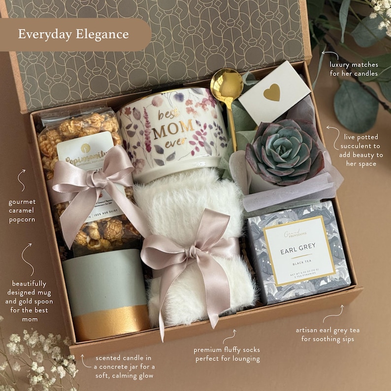 May include: A gift box filled with gourmet caramel popcorn, a floral "Best Mom Ever" mug, a gold spoon, a succulent, Earl Grey tea, a candle, fluffy socks, and matches. The box is tied with a satin ribbon.