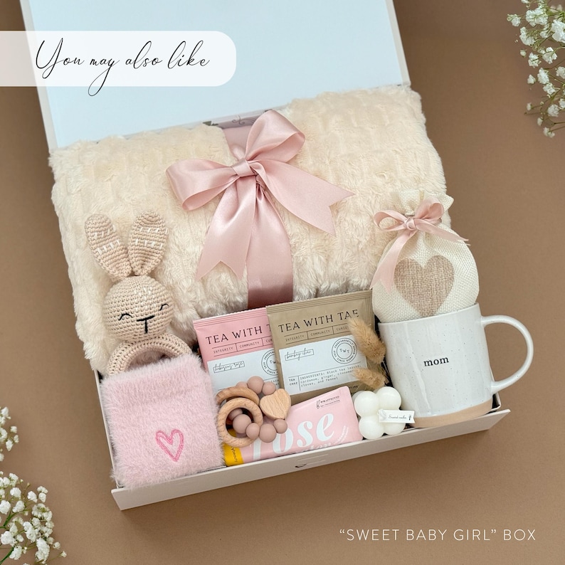 May include: A gift box for a baby girl, filled with a plush blanket, a crocheted bunny, a mug with "mom" printed on it, tea bags, a wooden teether, a pink heart-shaped item, and a candle. The box is white with a light pink bow.