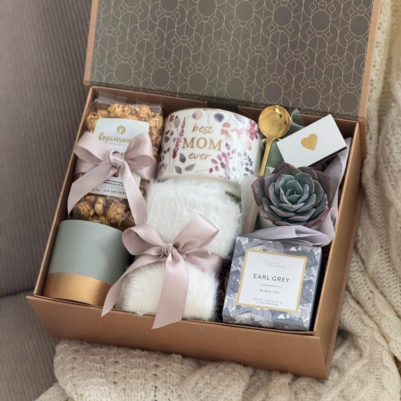 May include: A gift box with various items, including a mug that says "best MOM ever", popcorn, a succulent, a gold spoon, a small card, a candle, a tea box, and a fluffy white item, all tied with pink ribbons.