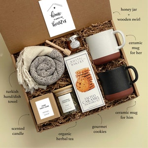 May include: A gift box filled with various items, including a Turkish hand towel, a scented candle, a jar of organic herbal tea, gourmet cookies, a honey jar with a wooden swirl, and two ceramic mugs. The box is decorated with a card that says "home is where the heart is".