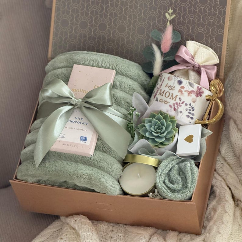 May include: A gift box with a sage green blanket tied with a satin ribbon, a mug that says "Best Mom Ever", a chocolate bar, a succulent, a candle, and a small card with a gold heart. The box is lined with patterned paper.