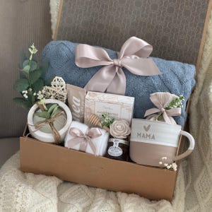 May include: A gift box filled with a blue blanket, a mug with "MAMA" printed on it, a small jar of honey, a bar of soap, a small plant, and other items. The box is tied with a light pink ribbon.