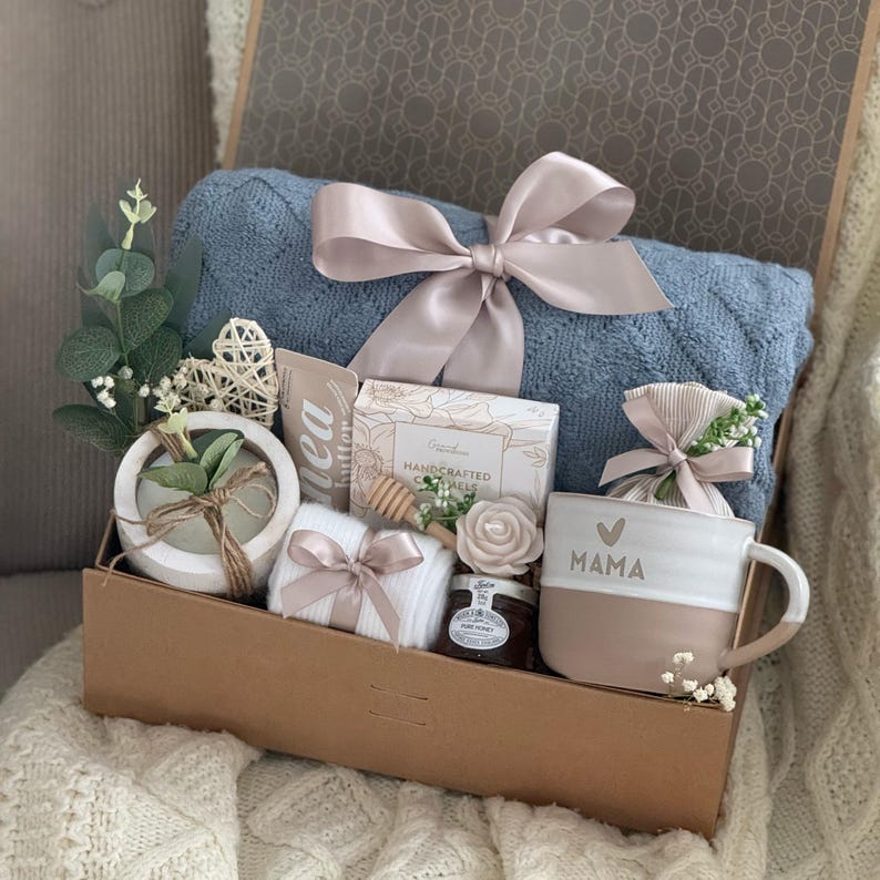 May include: A gift box filled with a blue blanket, a mug with "MAMA" printed on it, a small jar of honey, a bar of soap, a small plant, and other items. The box is tied with a light pink ribbon.