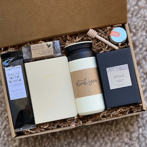 May include: A gift box filled with brown packing paper and various items, including a black bag of coffee labeled "Coffee early bird blend", a white notebook with gold lettering that says "Notes", a white travel mug with a brown label that says "thank you", a small black box with a white label that says "HYGGE Scented Candle Vanilla Bean", a small jar of honey with a wooden honey dipper, and a small black box with a white label that says "twenty-four blackbirds".