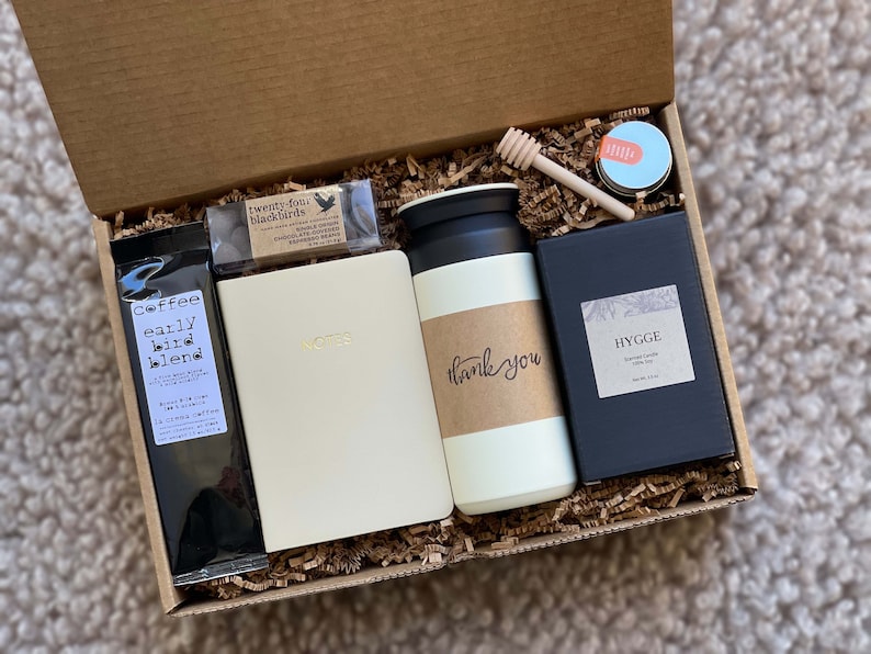 May include: A gift box filled with brown packing paper and various items, including a black bag of coffee labeled "Coffee early bird blend", a white notebook with gold lettering that says "Notes", a white travel mug with a brown label that says "thank you", a small black box with a white label that says "HYGGE Scented Candle Vanilla Bean", a small jar of honey with a wooden honey dipper, and a small black box with a white label that says "twenty-four blackbirds".