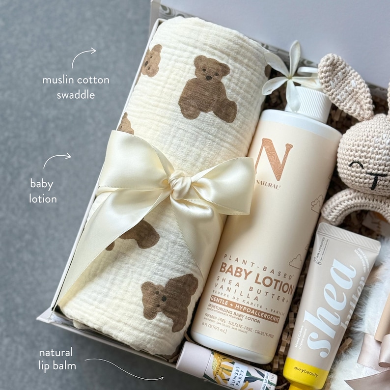 May include: A baby gift set featuring a muslin cotton swaddle with teddy bear prints, a bottle of plant-based baby lotion, a tube of shea lotion, and a natural lip balm. The set is tied with a cream-colored ribbon and includes a crocheted bunny.