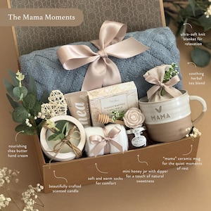 May include: A gift box with a blue knit blanket, a ceramic mug with "MAMA" on it, a scented candle, shea butter hand cream, soft socks, a honey jar, and a tea blend. The box is tied with a satin ribbon.