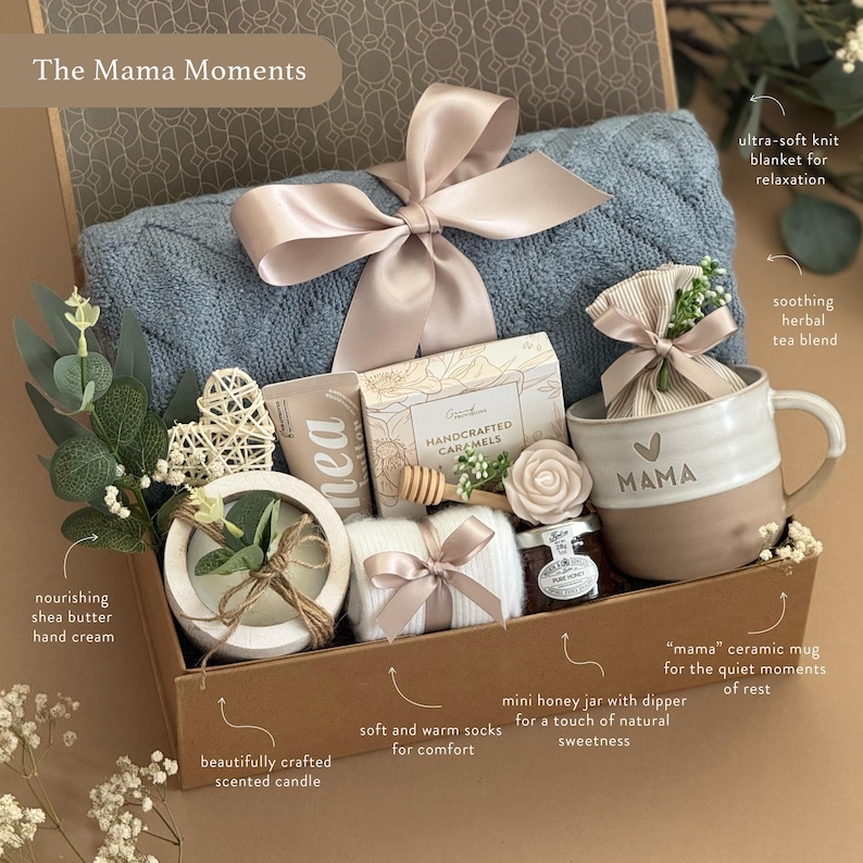May include: A gift box with a blue knit blanket, a ceramic mug with "MAMA" on it, a scented candle, shea butter hand cream, soft socks, a honey jar, and a tea blend. The box is tied with a satin ribbon.