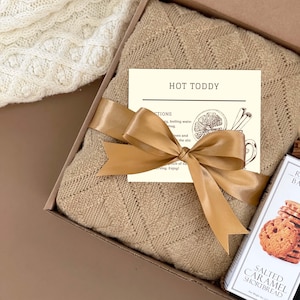 Puede incluir: Una caja de regalo llena de una acogedora manta de punto marr&oacute;n, un paquete de galletas de mantequilla salada con caramelo y una tarjeta de receta para un ponche caliente. La caja est&aacute; atada con una cinta dorada.