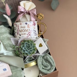 May include: A gift box filled with a floral-decorated mug that reads "best MOM ever", a gold spoon, a succulent, a candle, a small gift box with a gold heart, and a green blanket. The box is tied with a pink bow.