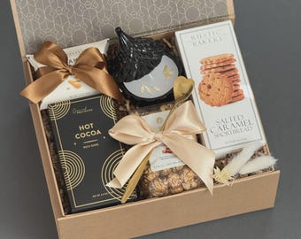 May include: A gift box filled with gourmet treats. Includes a black decorative item, a box of salted caramel shortbread, hot cocoa, and other snacks, all tied with gold satin bows. The box has a patterned lid.