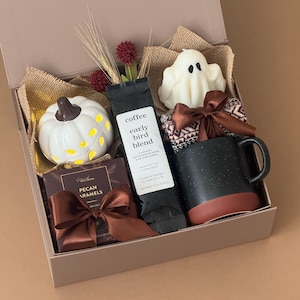 May include: A gift box containing a white ceramic pumpkin, a bag of Early Bird Blend coffee, a white ghost-shaped candle, a black and terracotta mug with a brown ribbon, and a box of Pecan Caramels with a brown ribbon, all nestled in burlap.