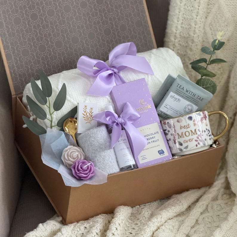 May include: A gift box filled with a white blanket, a mug with "best MOM ever" text, a chocolate bar, tea, a small towel, and other items. The box is tied with a lavender ribbon and decorated with a gold spoon and floral accents.