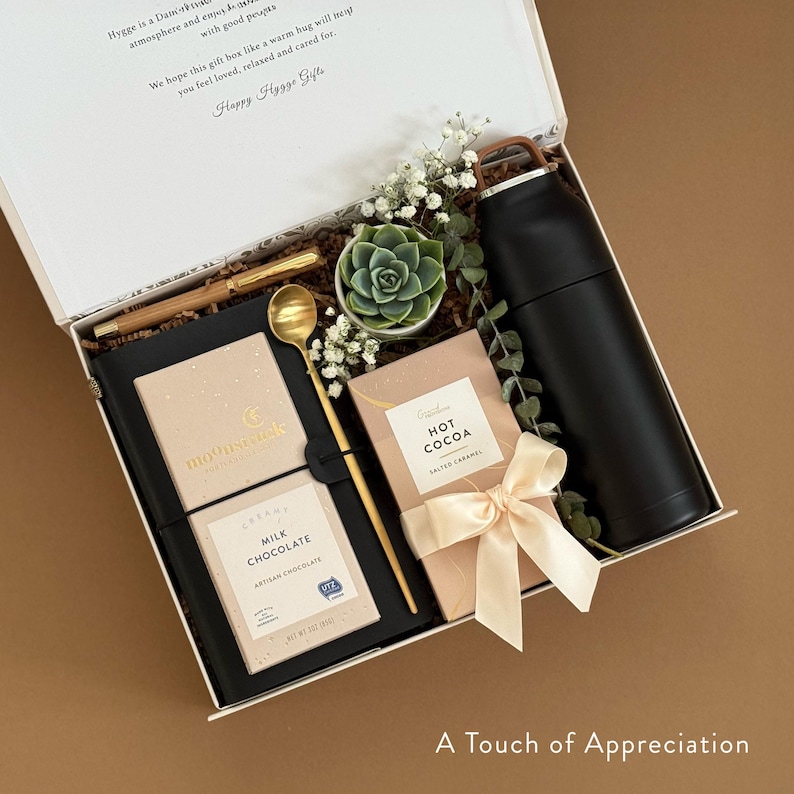 May include: A white gift box with a black lid, filled with a variety of items, including a notebook, a gold spoon, a small potted succulent, a black travel mug, and two boxes of chocolate. The box is labeled "A Touch of Appreciation".