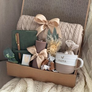 May include: A gift box filled with a beige knitted blanket, a green journal with a pen, a brown candle with a bow, a white and brown mug with "MAMA" text, and other decorative items. The box is tied with a beige ribbon.