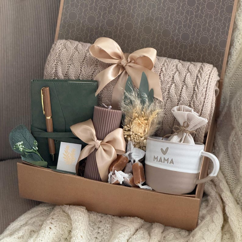 May include: A gift box filled with a beige knitted blanket, a green journal with a pen, a brown candle with a bow, a white and brown mug with "MAMA" text, and other decorative items. The box is tied with a beige ribbon.