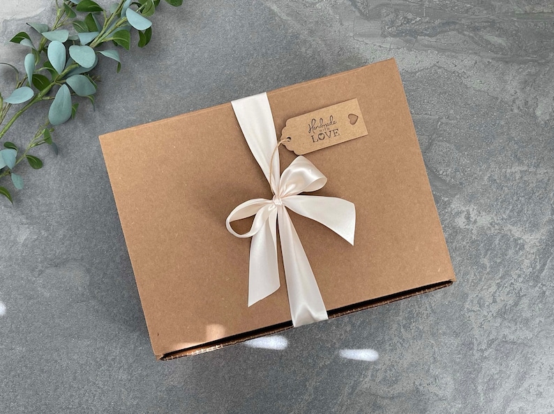 May include: A brown cardboard box with a white satin ribbon tied in a bow. The box has a small brown tag attached to the ribbon with the text "Handmade with LOVE" and a small heart.