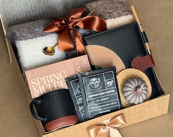 May include: A gift box filled with a black speckled mug, a blanket, a journal, coffee, a candle, and a spoon. The box is tied with a brown satin ribbon. The text "SPRING MULBE" is visible on a box.