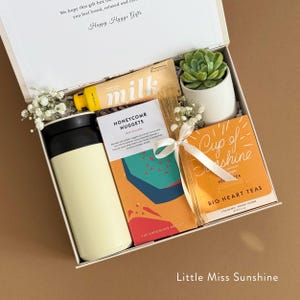 May include: A white gift box filled with various items, including a yellow tube of milk chocolate, a small potted succulent, a travel mug, a box of honeycomb nuggets, and a box of herbal tea. The box is labeled "Happy Hygge Gifts" and "Little Miss Sunshine".