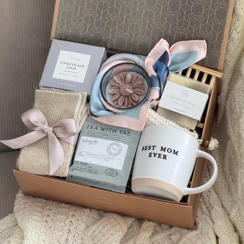 May include: A gift box filled with self-care items. Includes a mug that says "BEST MOM EVER", a candle, tea, socks, chocolate, a scarf, and soap. The box is brown with a decorative lid.