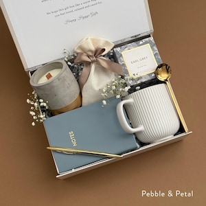 May include: A white gift box filled with a white mug, a gray concrete candle, a small white bag with a brown ribbon, a blue notebook with gold lettering that says "NOTES", a gold pen, a small tin of Earl Grey tea, and a gold spoon. The box is on a brown surface and has the text "Pebble & Petal" printed on the bottom.
