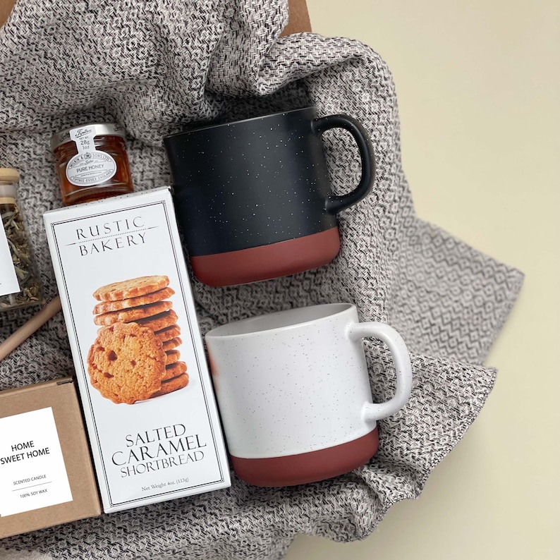 May include: A gift basket with a black and white speckled mug, a white mug with black speckles, a box of salted caramel shortbread cookies, a jar of honey, and a scented candle. The basket is lined with a gray and white patterned blanket.