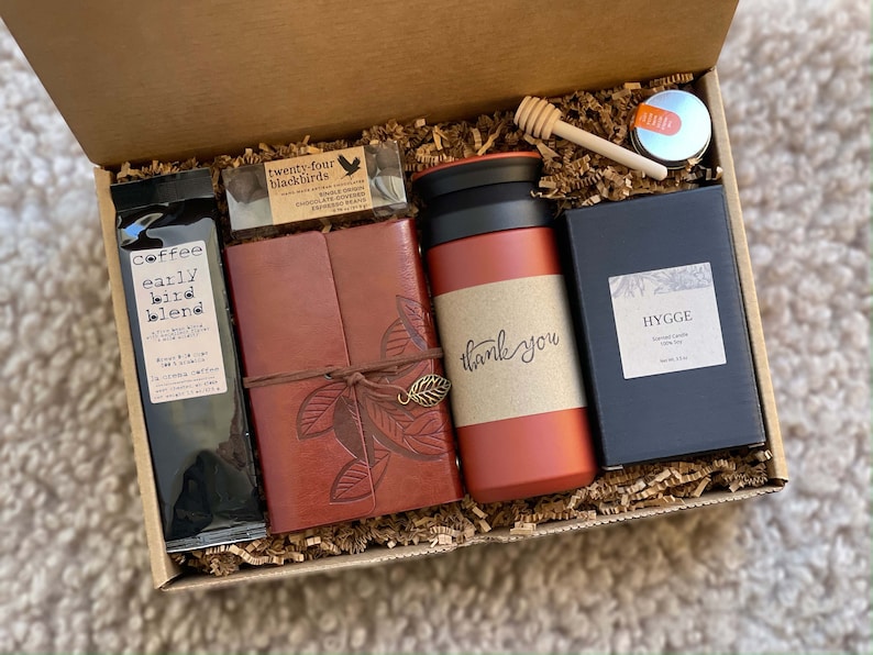 May include: A gift box filled with various items, including a brown leather journal with a leaf design, a red travel mug with "thank you" written on it, a bag of coffee labeled "Coffee Early Bird Blend", a small bar of chocolate labeled "Twenty-Four Blackbirds", a small jar of honey with a wooden honey dipper, and a black box with a candle labeled "Hygge Scented Candle Soy Wax".