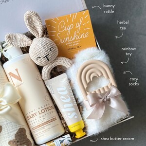 May include: A gift box containing baby products. Includes a crochet bunny rattle, a bottle of baby lotion, a tube of shea butter cream, a box of herbal tea, a rainbow toy, and cozy socks. The lotion bottle is labeled "N NATURAL".