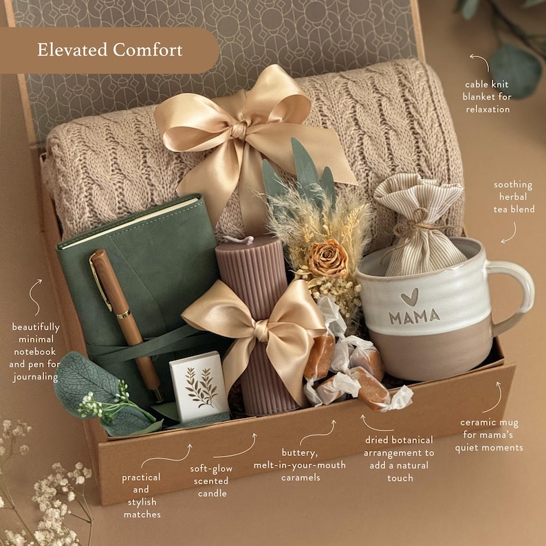 May include: A gift box with a beige cable knit blanket, a green notebook and pen, a candle, a mug with "MAMA" on it, caramels, and a dried botanical arrangement. The box is tied with a gold ribbon.