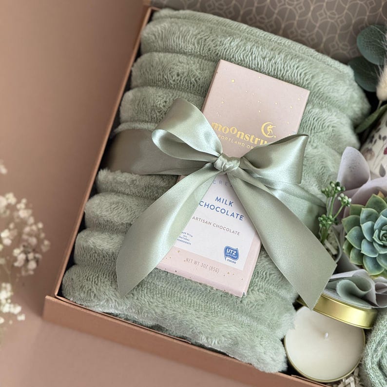 May include: A gift box with a sage green plush blanket, a pink box of milk chocolate, and a small candle. The blanket is tied with a matching satin ribbon. The chocolate box reads "MILK CHOCOLATE ARTISAN CHOCOLATE".