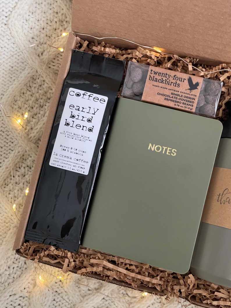 May include: A gift box filled with brown paper shreds contains a bag of coffee labeled "coffee early bird blend", a small box of chocolate covered espresso beans labeled "twenty-four blackbirds", a green notebook with the word "NOTES" in gold lettering, and a brown paper wrapped item with the word "thank" visible.