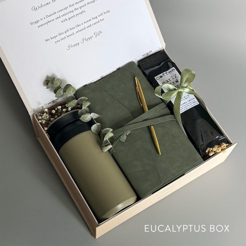 May include: A green gift box with a green journal, a gold pen, a green travel mug, and a bag of coffee. The box is tied with a green ribbon and has the text "EUCALYPTUS BOX" printed on the bottom. The box also includes a note that reads "Welcome Hygge is a Danish concept that means atmosphere and enjoying the good things with good people. We hope this gift box like a warm hug will help you feel loved, relaxed and cared for. Happy Hygge Gifts"