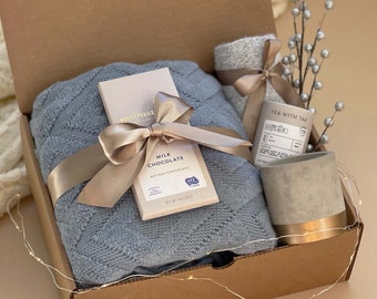 Può includere: Una scatola regalo di cartone marrone piena di una coperta di maglia blu, una barretta di cioccolato al latte, un piccolo portacandele in cemento grigio con bordo dorato e una piccola scatola di bustine di tè grigia e bianca con il testo "Tea with Tae" sul davanti.